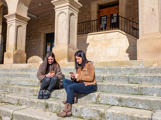 Two pretty women sitting on the stairs looking at their cell phones.