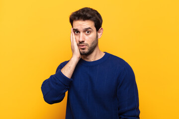 young handsome man feeling shocked and astonished holding face to hand in disbelief with mouth wide...