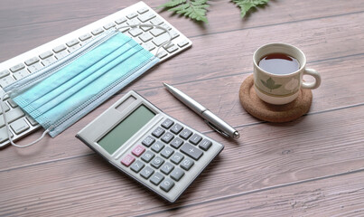 Wood table with keyboard, face mask, coffee, pen, calculator and green plants.