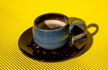 Delicious freshly brewed coffee in a blue cup on a brown plate with coffee beans on a yellow table.