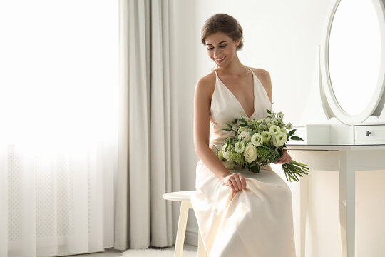 Young bride in wedding dress with beautiful bouquet near mirror indoors
