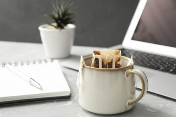 Cup with drip coffee bag, laptop and notebook on light grey marble table, closeup