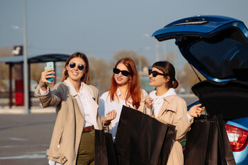 Obraz premium Young women at the car with shopping bags. Girls take selfie