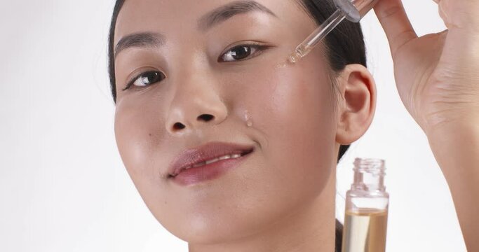 Close up portrait of young asian woman applying serum on her cheek, skin care, white studio background