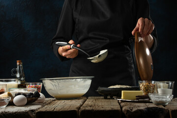Pastry chef in black uniform pours dough into waffle maker machine for preparing sweet waffle on rustic wooden table with ingredients on dark blue. Frozen motion. Dessert from recipe book.