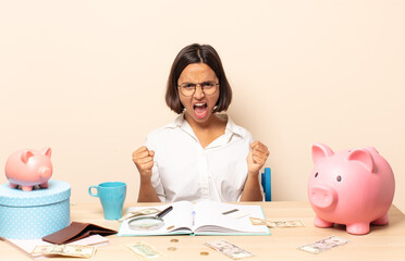 young latin woman shouting aggressively with an angry expression or with fists clenched celebrating...