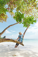 Asian woman sitting on the tree and enjoying with beautiful sea nature in her vacation. Summer holidays and family travel concept. Travelling in Thailand.