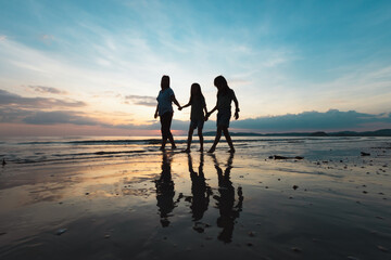 Silhouette of asian mother and two daughters holding hand and walking on the beach together at the sunset time with beautiful sea and sky. Family enjoy with nature concept.