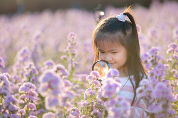 Cute asian little child girl holding magnifier looking on flower with curiously and enjoying with beautiful flower in the flower garden.