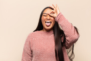 young latin woman smiling happily with funny face, joking and looking through peephole, spying on...