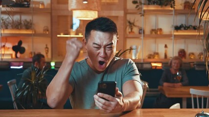Excited cheerful chinese man using smartphone winning online game money prize celebrating his victory in cafe. Carefree lifestyle. Lucky people. Emotions.