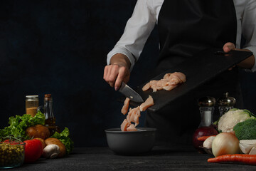 The professional chef in black apron pours chicken fillet into frying pan for cooking soup on dark blue background. Backstage of preparing meal. Healthy dish. Food concept. Frozen motion. Cookbook.