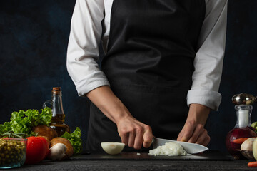 Professional chef in black apron cuts with knife onion on chopped board for cooking soup on dark blue background. Backstage of preparing meal. Healthy dish. Food concept. Frozen motion. Cookbook.