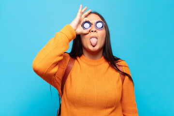 young latin woman smiling happily with funny face, joking and looking through peephole, spying on...