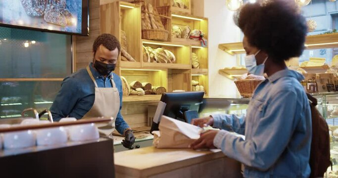 Side view of African American male seller in face mask selling baked fresh bread in bakery shop to pretty female client. Woman paying with credit card buying baking in bakehouse. Shopping concept