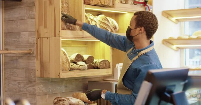 Side view. African American young man worker wearing face mask and apron standing in bakery shop and putting fresh baked bread on shelves. Male bakehouse owner working alone in small coffee shop