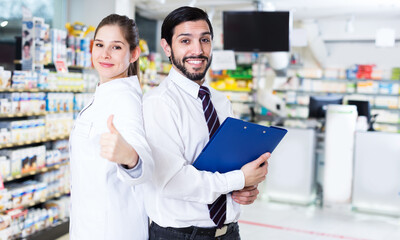 Female pharmacist is standing with man expert with folder in pharmacy