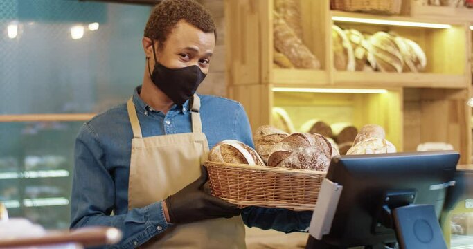 Close up of handsome happy young African American man bakery worker in face mask standing in bakehouse and holding basket with fresh baked bread. Bakery owner looking at camera and smiling