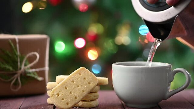 Making instant coffee. Scottish shortbread cookies and wrapped Christmas gift on table. Christmas tree at background.