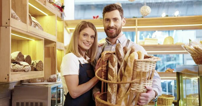 Portrait of joyful lovely Caucasian young family married couple of bakers in good mood holding basket with fresh bread standing in small own bakery shop and smiling at camera. Family business concept