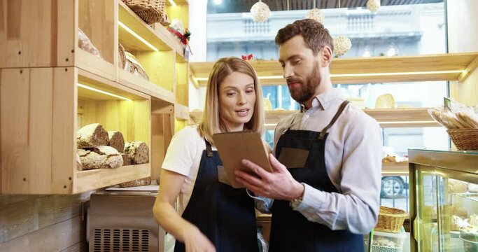 Portrait of happy Caucasian married couple in aprons standing in bakehouse talking and browsing on device. Man in small own bakery shop with co-worker tapping and typing on tablet at work Bake concept