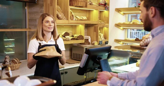 Rear of Caucasian male customer buying fresh bread in bakery shop and paying with credit card. Pretty female seller in apron selling baking in bakehouse. Small business. Shopping concept