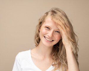 Fototapeta premium Beautiful young girl laughing. Pretty blonde with curly hair in white t shirt against beige wall smiling looking at camera and covering face with hand
