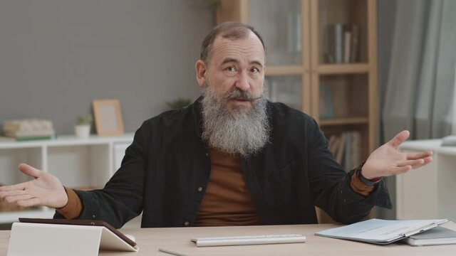 Chest Up POV Of Senior Caucasian Man With Heavy Silver Beard Sitting By Desk At Home Office, Looking On Camera, Speaking Emotionally To Invisible Partner On Video Call