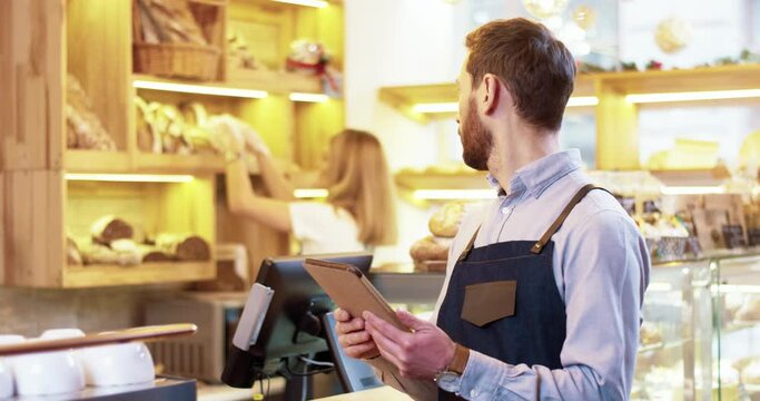 Close up of handsome bearded young Caucasian man seller in apron standing in bakery, tapping and typing on tablet device while colleague works on background. Bakehouse concept