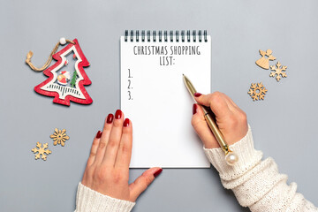 female hand holds pen, writing Christmas shopping list, gift ideas on white notepad on gray...