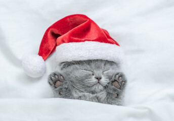 Funny kitten wearing red santa's hat sleeps under a white blanket on a bed. Top down view