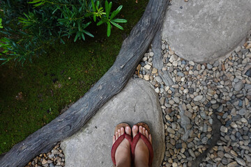 Asian young girl relax standing on grey stone while wearing Japanese shoes or wooden geta with red fabric in the traditional garden included tree bush, pebble and peaceful environment for rest mind
