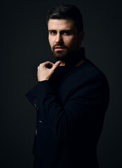 Thoughtful bearded businessman modern intellectual in jacket stands side and looking at camera holding hand at chin cover dark background. Stylish business casual style for men, portrait concept
