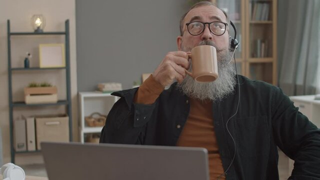 Medium Close-up Of Elderly Bearded Caucasian Man Wearing Glasses And Headset With Mic, Using Laptop For Video Call, Drinking From Mug, Leaning Back On Chair, Standing Up And Leaving