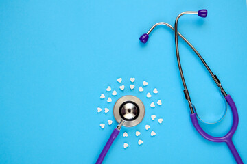 Stethoscope and white heart-shaped tablets in form of circle on a blue background with copy space. Concept of Valentine's day, medical Flatlay.