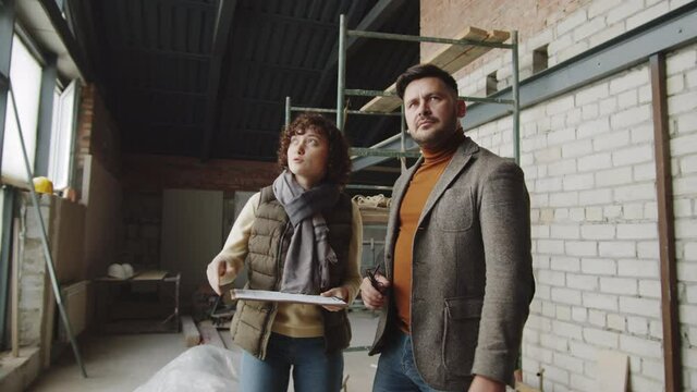 Young female architect walking with male client into unfinished apartment, showing him around and discussing construction plan on clipboard