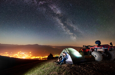 Happy couple man and woman tourists sitting in tourist tent on the top of mountain with atv quad motorbike, enjoying beautiful view of night sky full of stars, Milky way, luminous city on background © anatoliy_gleb