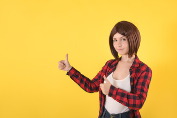 Portrait beautiful young woman standing in checked shirt and gave thumbs up: space for text, well signed sign and looking at the camera on yellow background