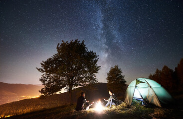 Young couple travellers resting at campfire near tourist tent, warming hands under amazing night sky full of stars and Milky way. On the background starry sky, mountains, big tree and luminous town