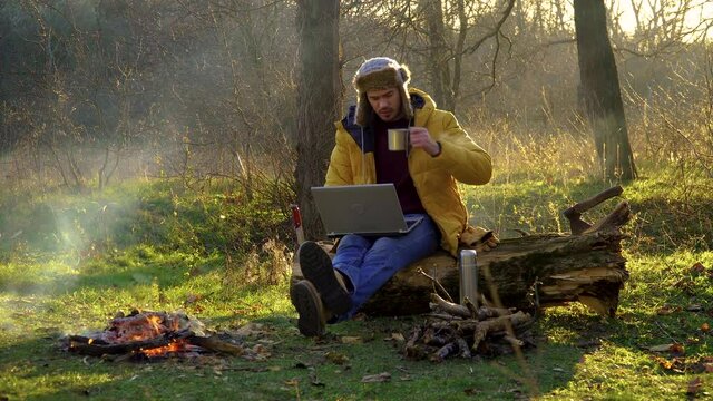 A young man works at a laptop near the fire in the forest. Freelance remote work concept. 4K