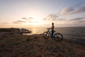 Naklejka na ściany i meble Woman with a bike in the nature / Morning view of a woman with an electric bike enjoys the view of sunrise at the rocky Black Sea coast.