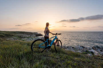 Woman with a bike in the nature / Morning view of a woman with an electric bike enjoys the view of sunrise at the rocky Black Sea coast.