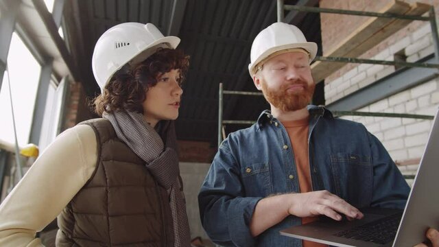 Waist up shot of professional male and female architects in hardhats standing together at construction site and discussing something on laptop