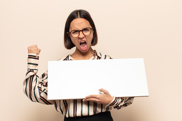 young latin woman shouting aggressively with an angry expression or with fists clenched celebrating...