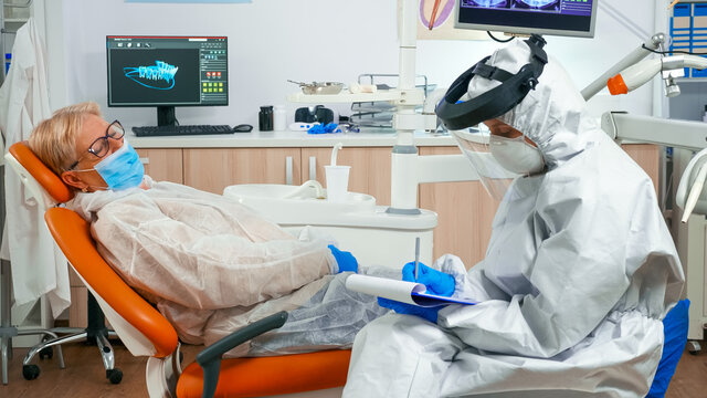 Nusre in protection suit measuring old woman temperature before dental examination during global pandemic. Assistant with face shield, mask, interrogating senior woman and taking notes on clipboard