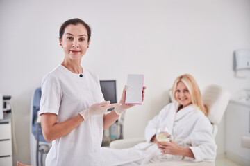 Beautiful professional cosmetologist standing near the patient with white notebook in room indoors