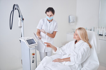 Obraz premium Happy smiling client looking to the list with programs of ultrasound modern machine in beauty center