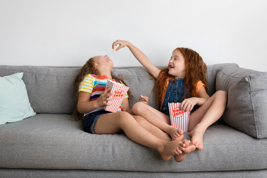 Two young girls sitting on couch feeding popcorn to each other