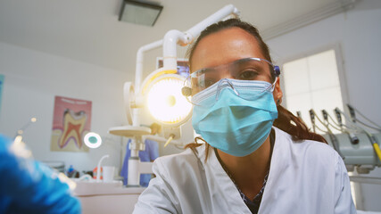 Fototapeta na wymiar Patient pov visiting dental clinic for surgery treating affected mass. Doctor and nurse working together in modern orthodontic office, lighting the lamp and examining person wearing protection mask.