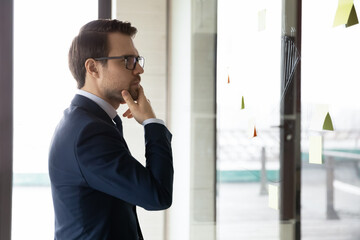 Thoughtful young Caucasian businessman look at glass wall work brainstorm using sticky pads developing business ideas. Pensive male worker do creative thinking in office. Startup, strategy concept.